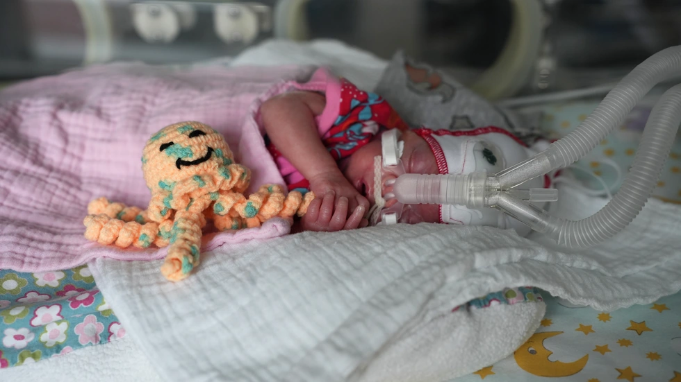 Emma liegt in ihrem Wärmebettchen, umgeben von Schläuchen, weichen Decken und gedämpftem Licht. Neben ihrem Gesicht sitzt eine kleine gehäkelte Krake. Emma liegt in ihrem Wärmebettchen, umgeben von Schläuchen, weichen Decken und gedämpftem Licht. Neben ihrem Gesicht sitzt eine kleine gehäkelte Krake.